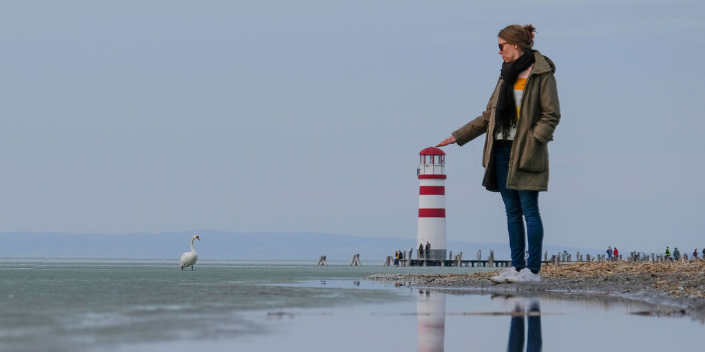Cómo fotografiar una ilusión óptica: mujer simulando tocar la parte superior de un faro gracias a la perspectiva en la playa.