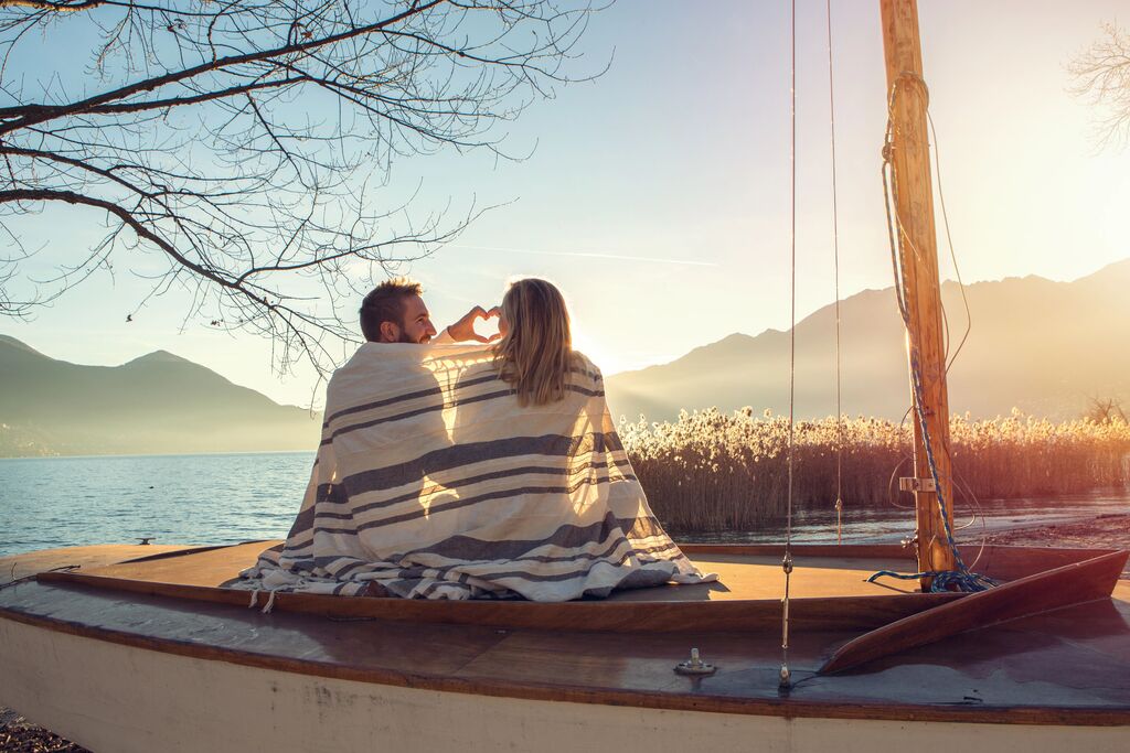 Una pareja está sentada en un barco, acurrucada en una manta, formando un corazón con sus manos. El sol brilla y el lago reluce.