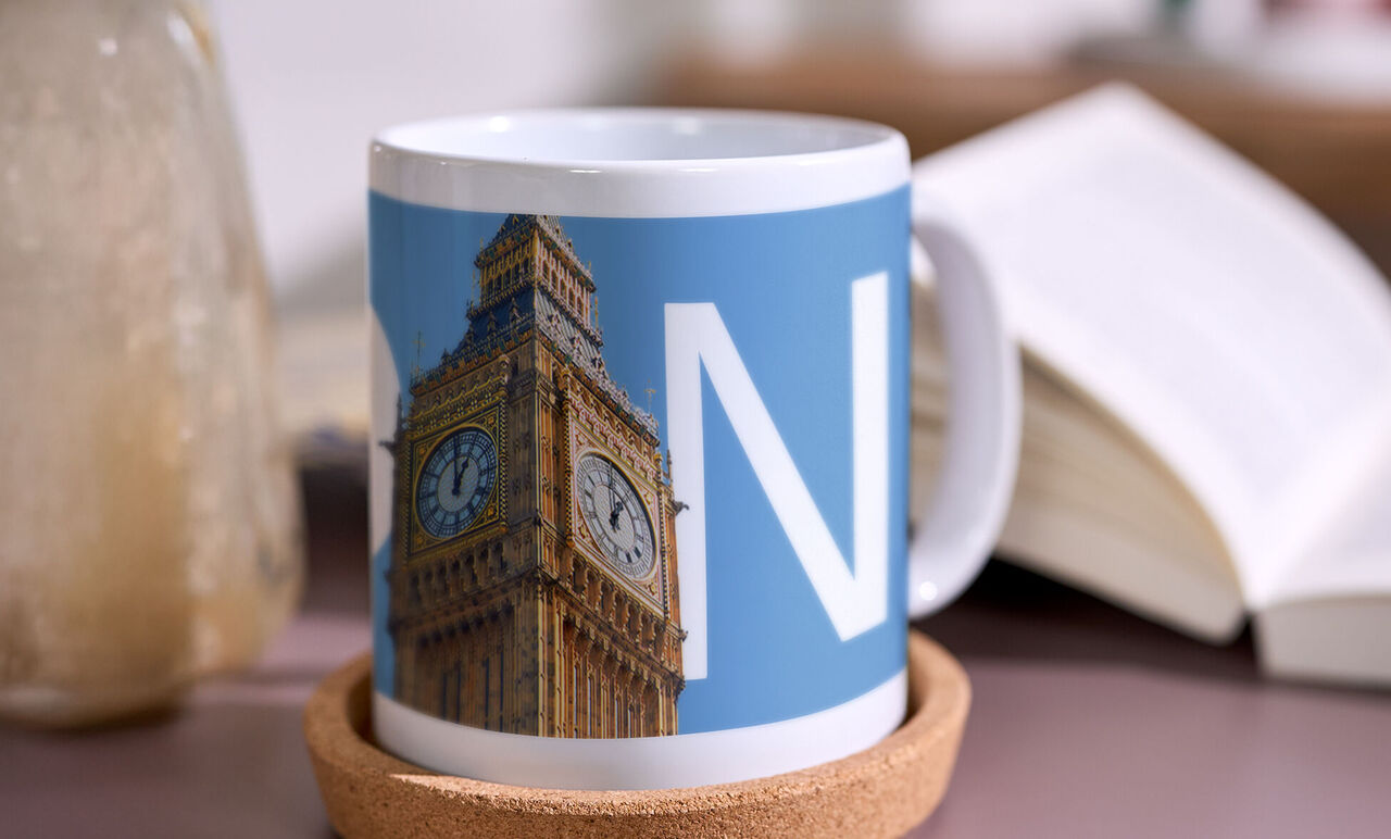Taza blanca personalizada con una ilustración del Big Ben y una gran letra “N” blanca sobre fondo azul, colocada junto a un libro abierto.
