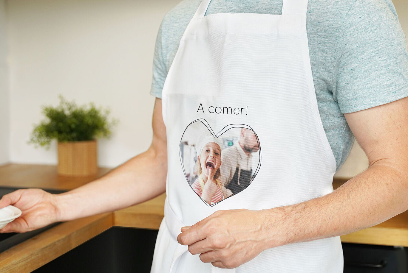 Una persona lleva un delantal blanco con un estampado de corazón y el texto '¡A comer!'. Fondo en una cocina moderna.