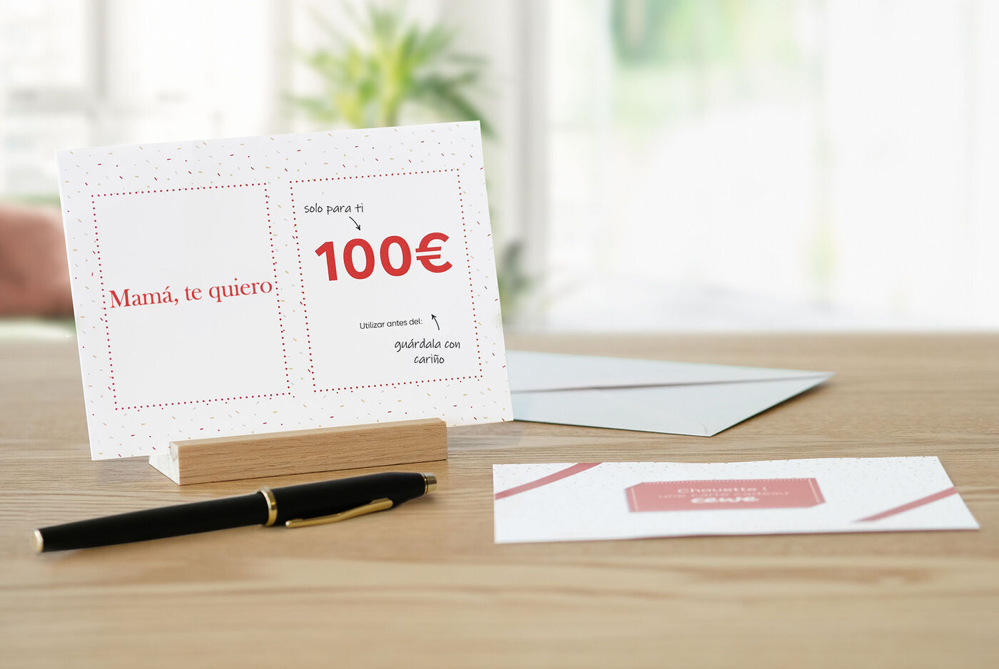 Una tarjeta de saludo CEWE con el texto 'Mamà, te quiero' y un código de 100€ sobre una mesa de madera.