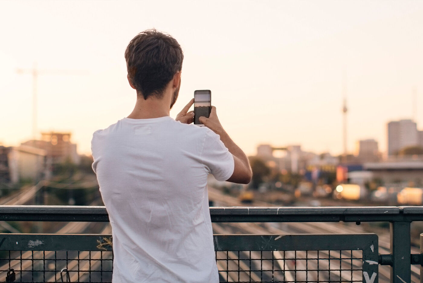 Hombre de espaldas fotografiando el skyline de una ciudad desde un puente al atardecer.