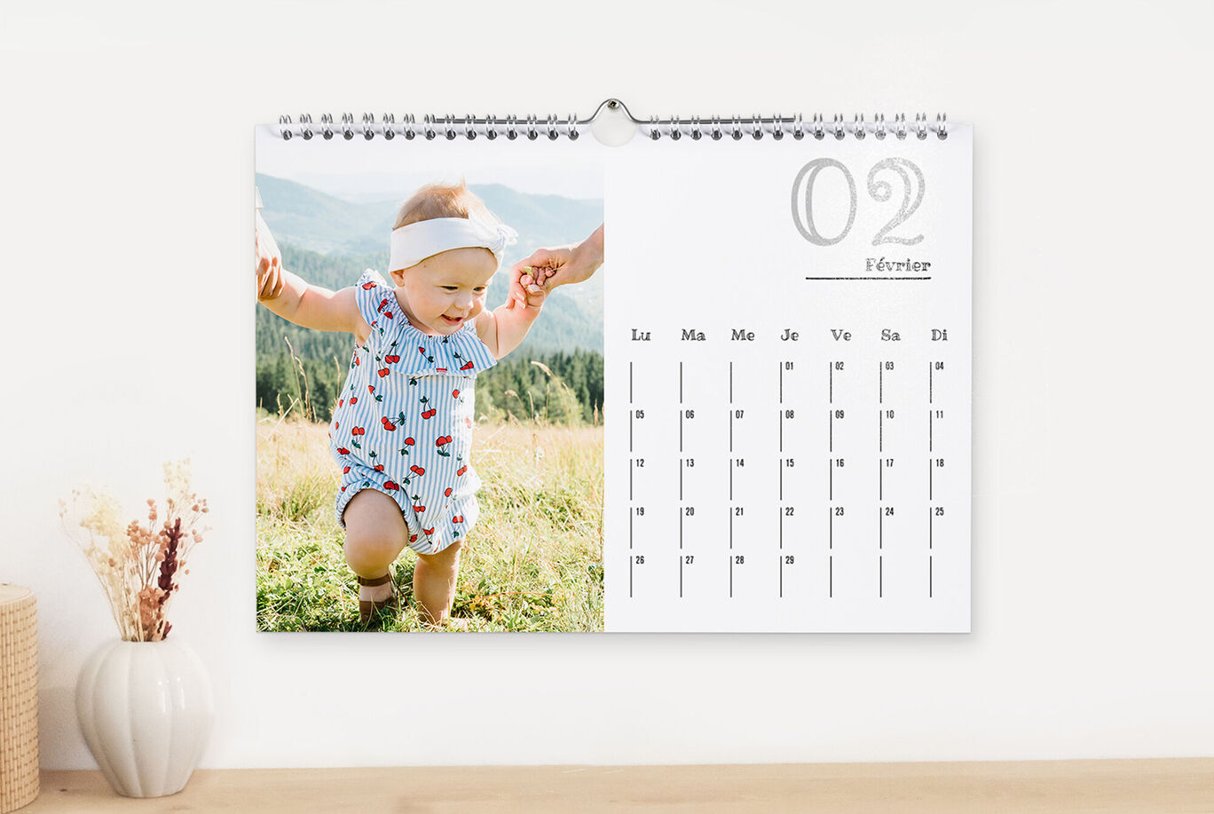 Un niño pequeño con una blusa colorida corre por un prado, sostenido por adultos. Al fondo hay colinas verdes.