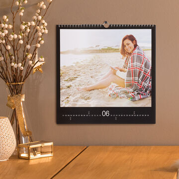 Una persona está en la playa mirando un calendario, rodeada de ramas decorativas y un objeto elegante en la mesa.