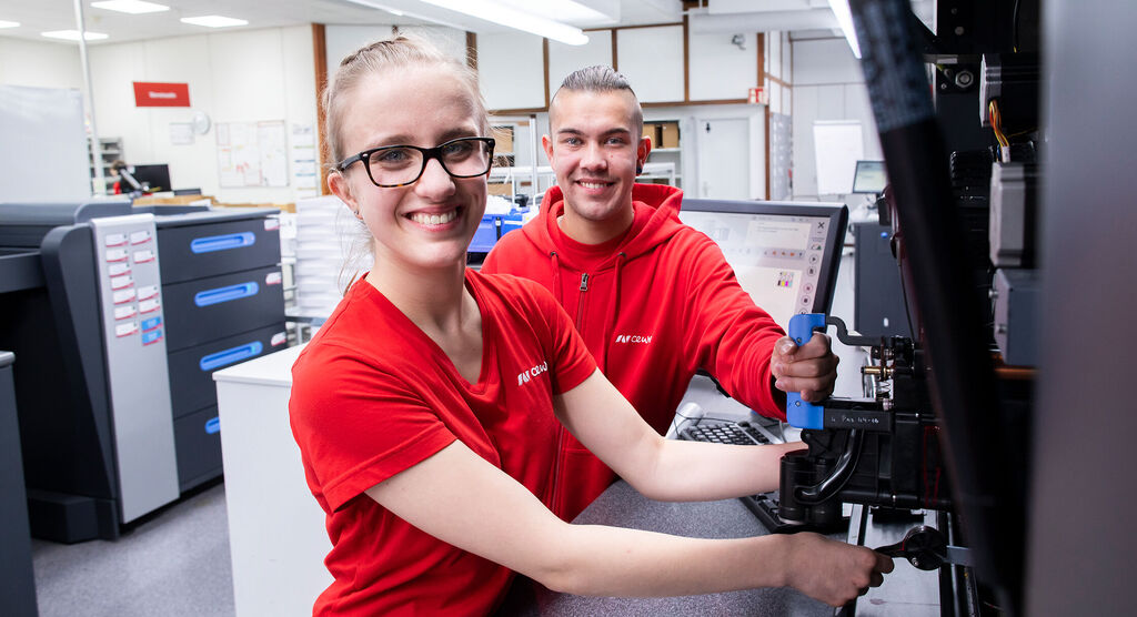 Dos empleados trabajan en una máquina en una oficina moderna, ambos llevan camisetas rojas.