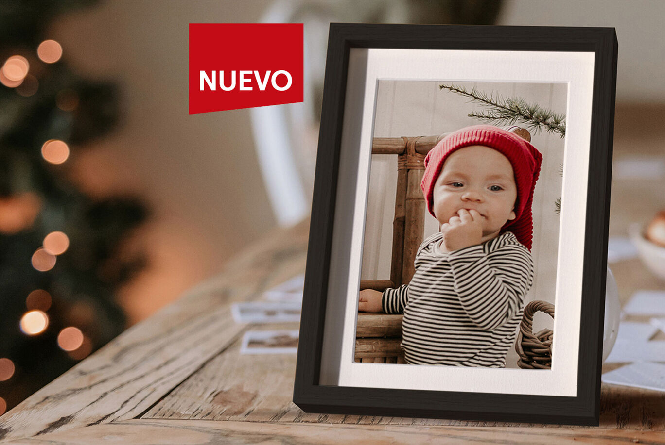 Un lindo bebé sentado en un marco de madera, sonríe usando un gorro rojo. Luces decorativas en el fondo crean una atmósfera cálida.