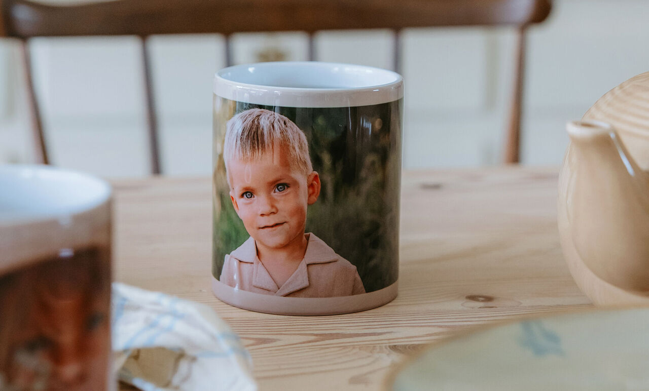 Taza personalizada sobre una mesa de madera con la fotografía de un niño rubio, un vaso desenfocado.