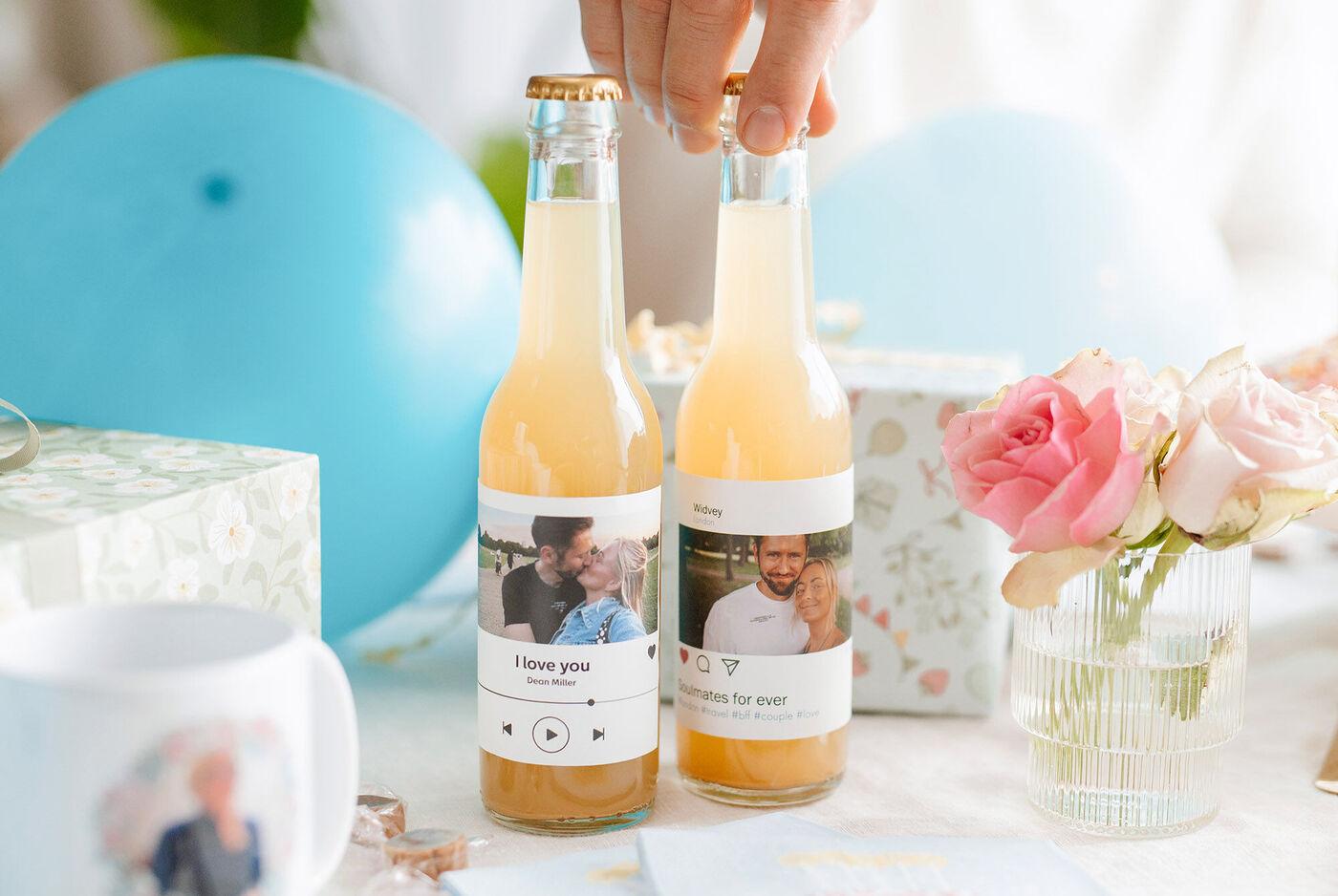 Dos botellas de zumo personalizadas con etiquetas de fotos, junto a globos azules, un ramo de rosas rosas y una taza blanca, sobre una mesa