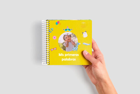 Una mano sostiene un libro amarillo con pegatinas coloridas y foto de un niño sonriente en la portada. Título "Mis primeras palabras".