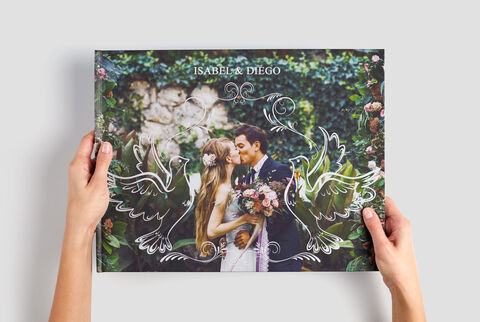 Una pareja sostiene un álbum CEWE de fotos de boda y está frente a una pared verde.