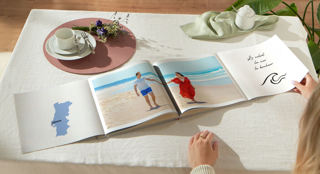 Una pareja mira un álbum fotográfico CEWE en la mesa, con fotos de sus vacaciones en la playa.