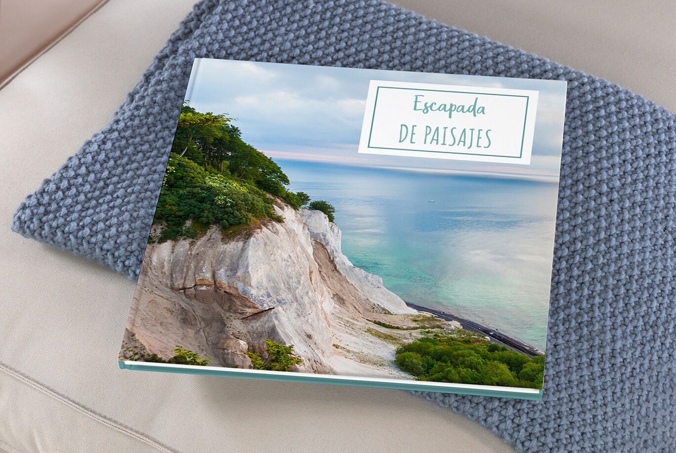 Un CEWE FOTOBUCH está sobre una manta de lana cómoda, con una hermosa imagen costera de fondo.