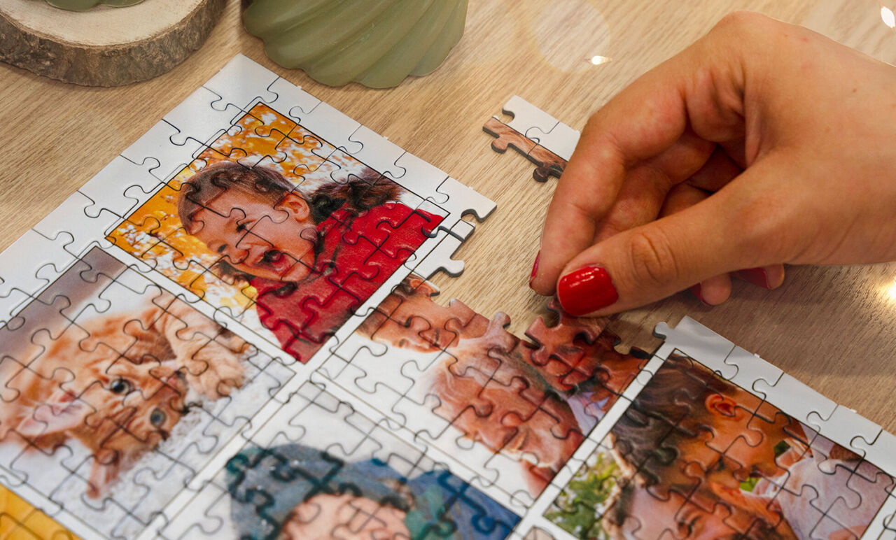Primer plano de una mano con uñas rojas colocando una pieza en un puzzle personalizado tipo collage que muestra fotos familiares.