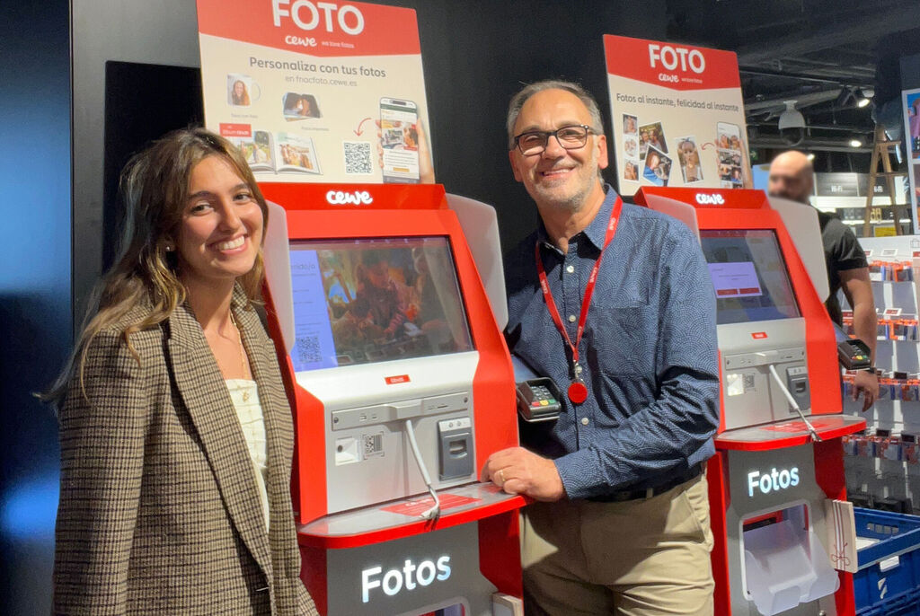 Dos personas junto a quioscos de impresión de fotos CEWE en un establecimiento, con expositores y productos de fondo.