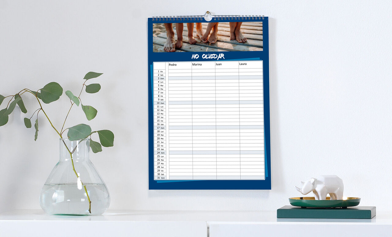 Un calendario fotográfico CEWE con un marco azul cuelga de la pared y muestra una lista con columnas para actividades y fechas.