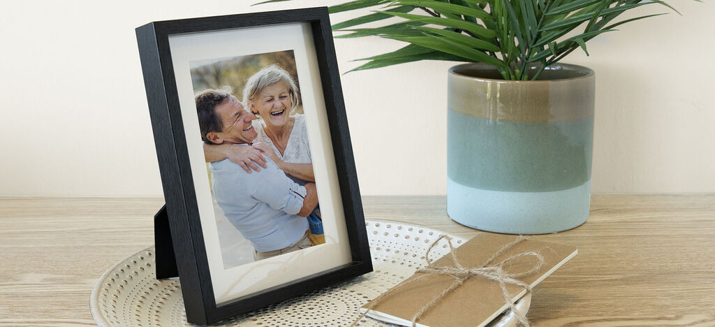 Una hermosa foto enmarcada de un abrazo amoroso sobre una mesa. Al lado hay un cuaderno y una planta.