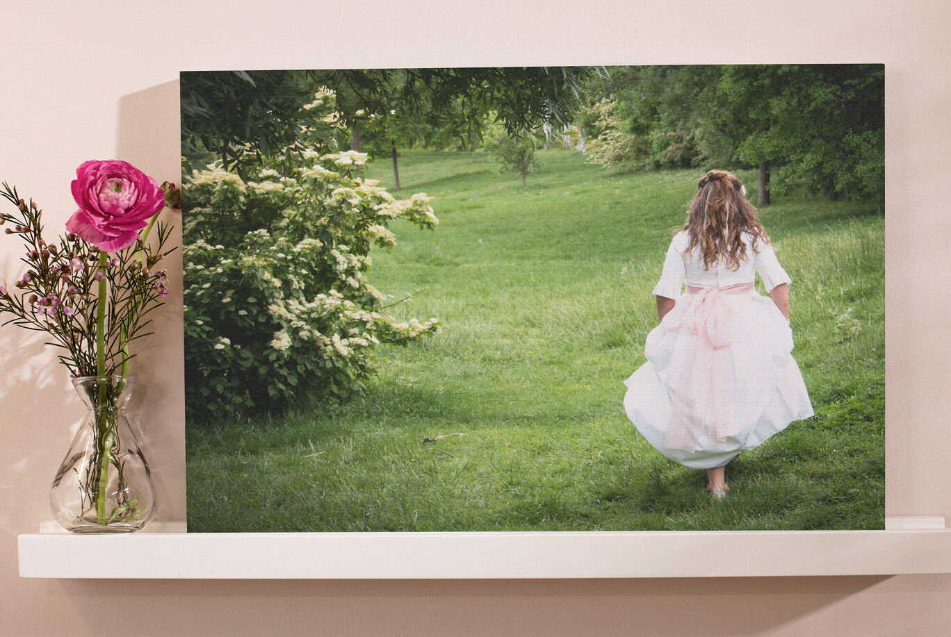 Una niña corre descalza en un campo verde. Lleva un vestido blanco y tiene cabello largo y castaño. Detrás florecen muchas plantas verdes. Hay un jarrón con una flor rosa en la mesa.