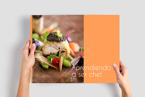 Una persona sostiene un álbum CEWE con un plato colorido que parece recién servido. La portada es naranja.