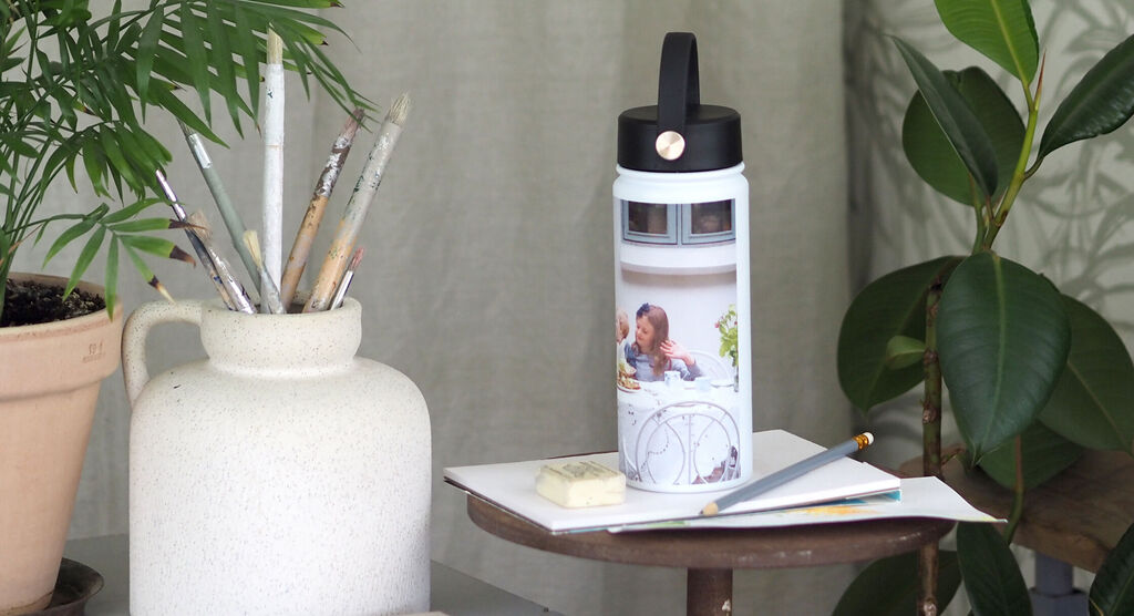 Persona bebiendo de una botella de agua personalizada, en un cuarto iluminado, rodeada de plantas y utensilios de dibujo