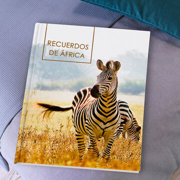 Un hermoso CEWE Fotobook con una portada de cebra sobre una superficie clara. Al fondo hay otras cebras y un paisaje suave.