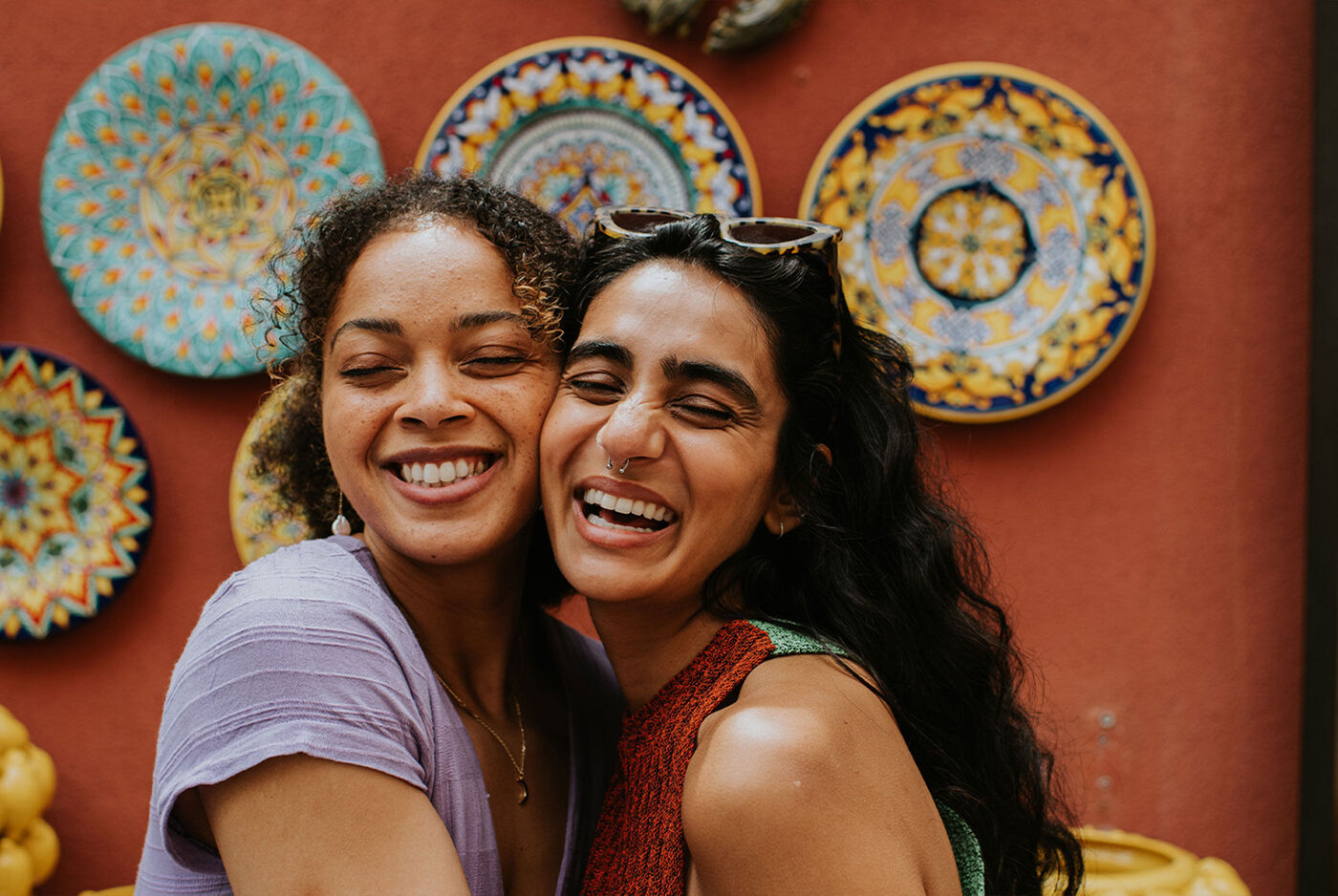 Dos mujeres jóvenes sonriendo y abrazándose frente a una pared roja decorada con platos de cerámica de colores.
