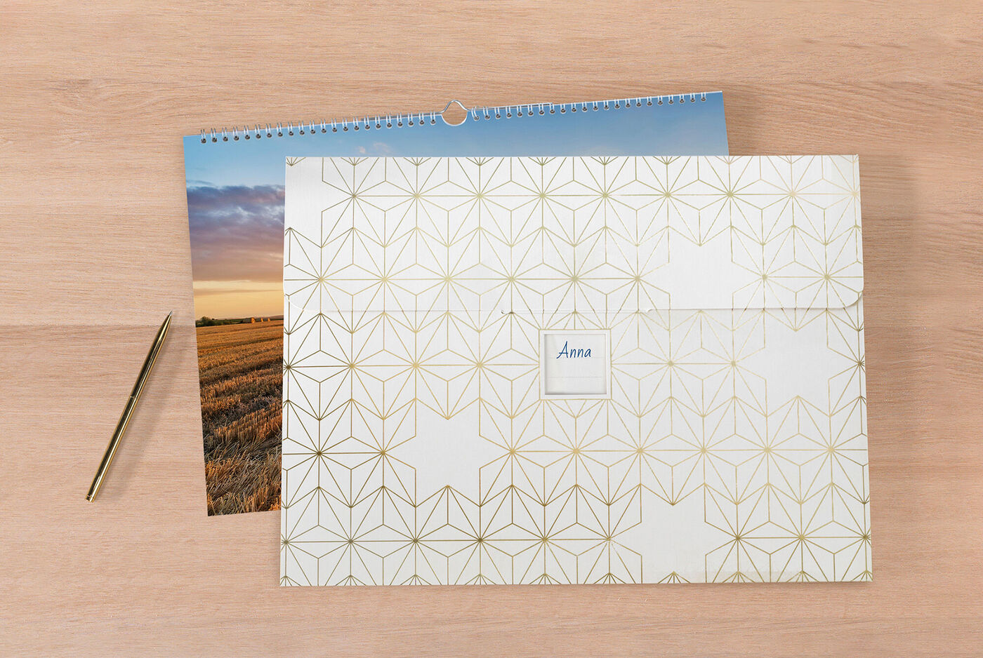 Un CEWE fotokalender bellamente diseñado sobre una mesa de madera, junto a un sobre decorativo con el nombre 'Anna' y un bolígrafo dorado.