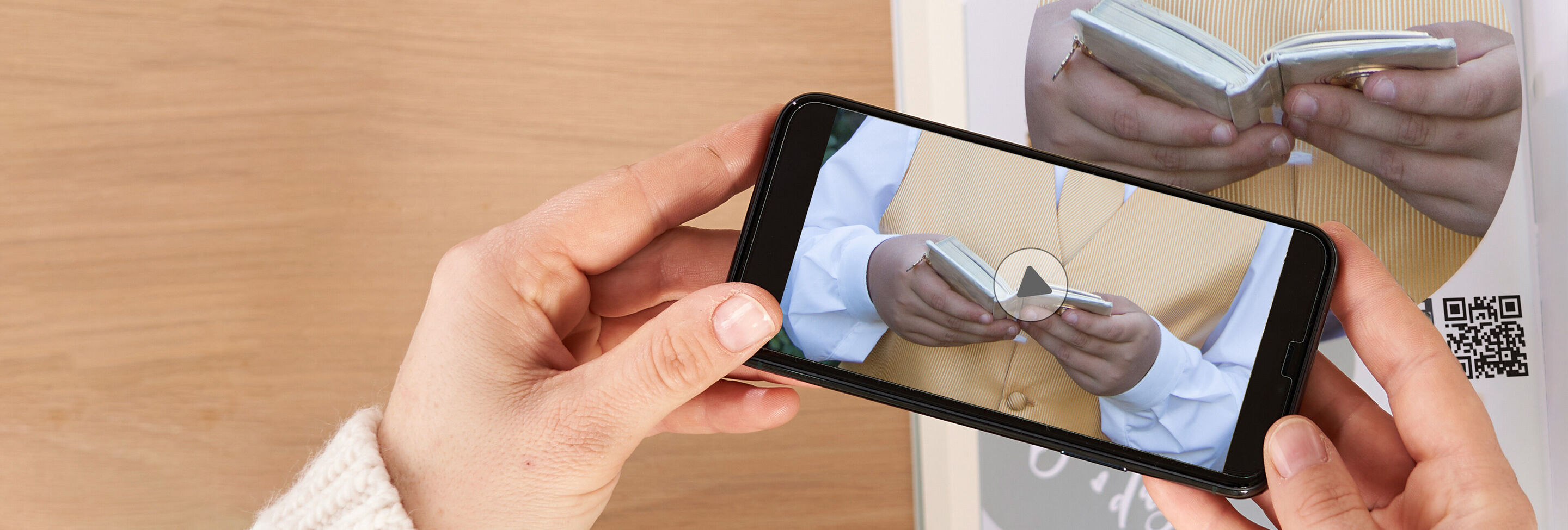 Manos sosteniendo un smartphone que muestra un libro de fotos enrollado, fondo de un folleto.