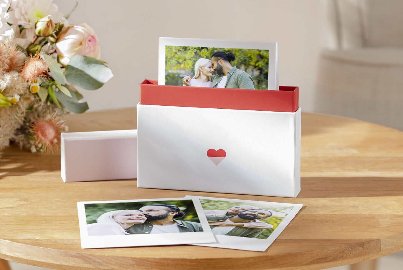 Una bonita caja blanca con un diseño de corazón rojo que contiene fotos. La caja está sobre una mesa de madera, junto hay algunas fotos de una familia feliz. En el fondo florecen hermosas flores.