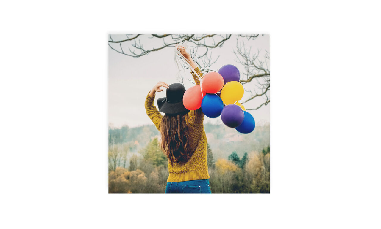 Mujer con cabello largo y castaño lleva un sombrero negro y sostiene globos de colores.