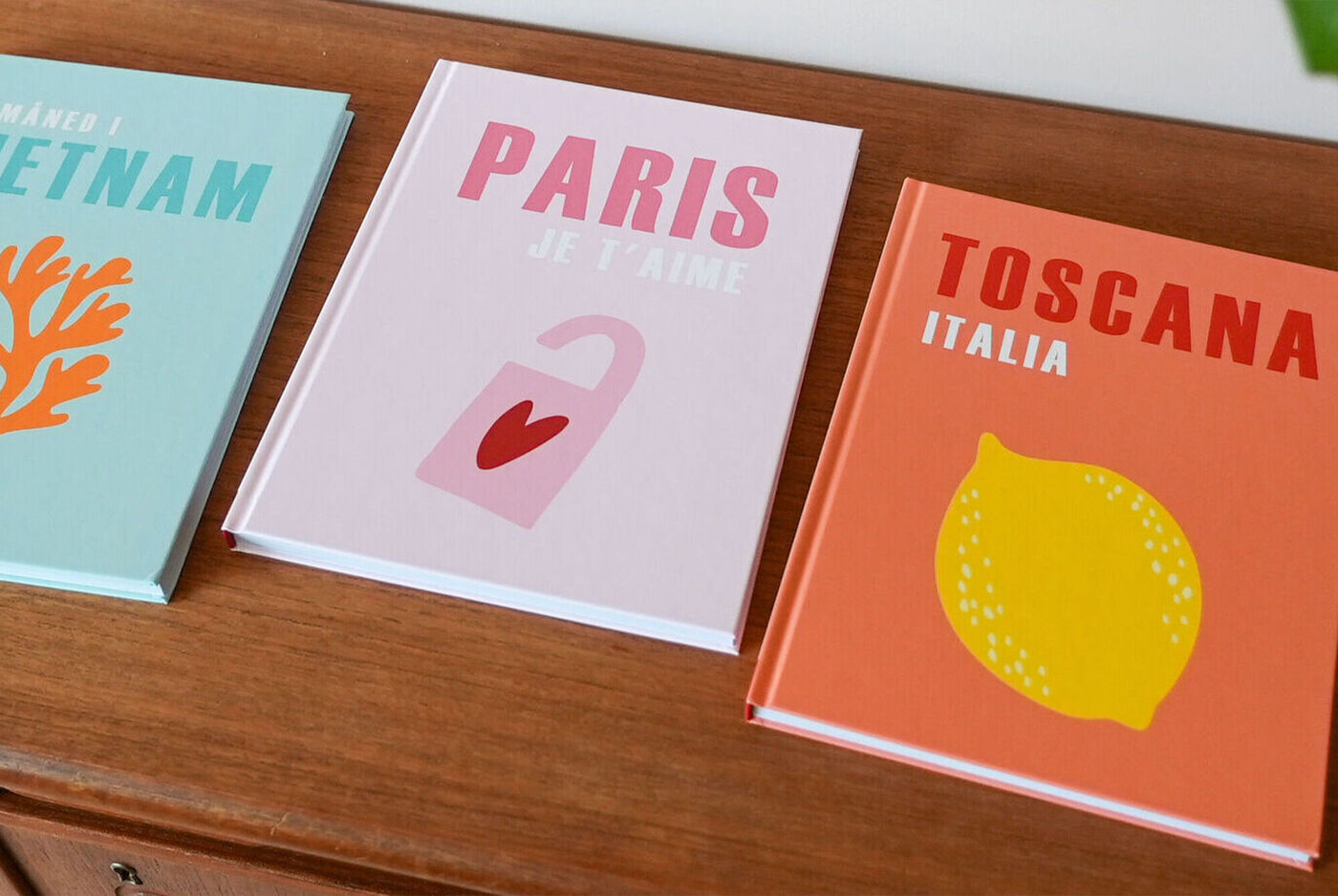 Tres álbums de viaje con portadas coloridas temáticas de Vietnam, París y Toscana sobre una mesa de madera.