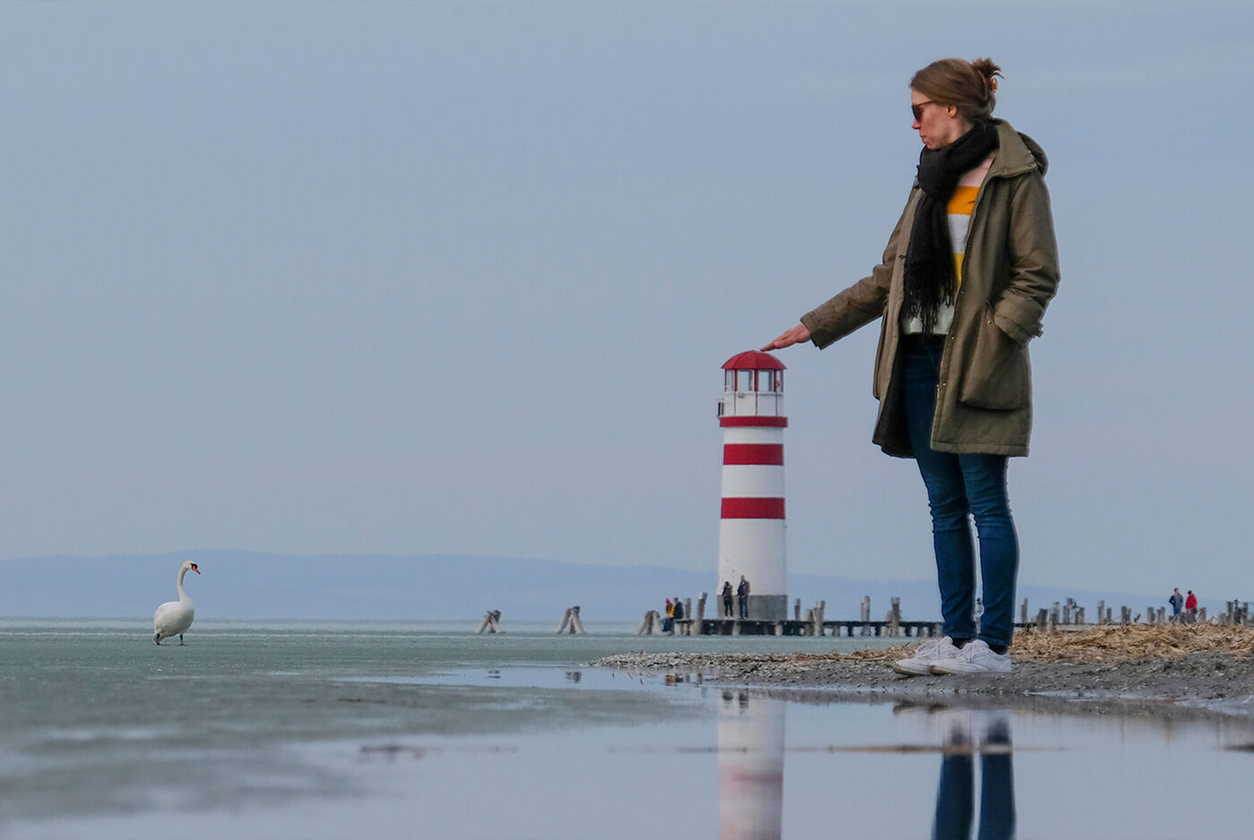 Mujer de pie frente a un faro rojo y blanco en la orilla del mar, con el reflejo en el agua y una gaviota al fondo