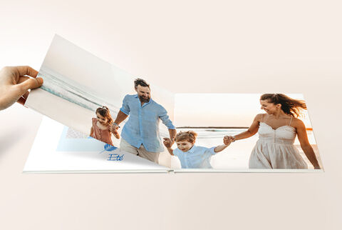 Una mano hojea un CEWE FOTOBUCH mostrando momentos del mar. Las páginas muestran familias felices de vacaciones.