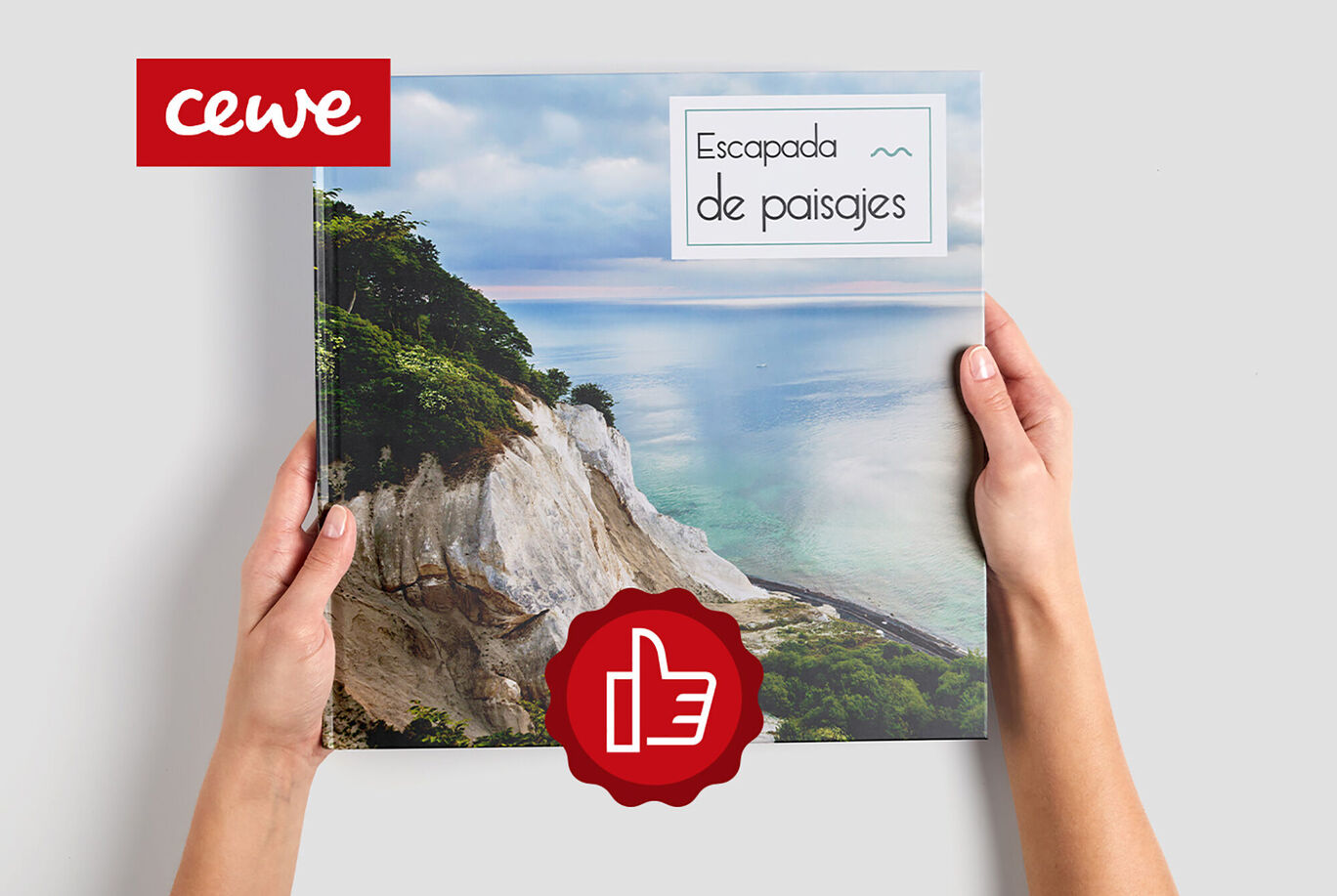 Dos manos sostienen un CEWE Fotolibro con fotografía de paisajes frente a un paisaje costero.