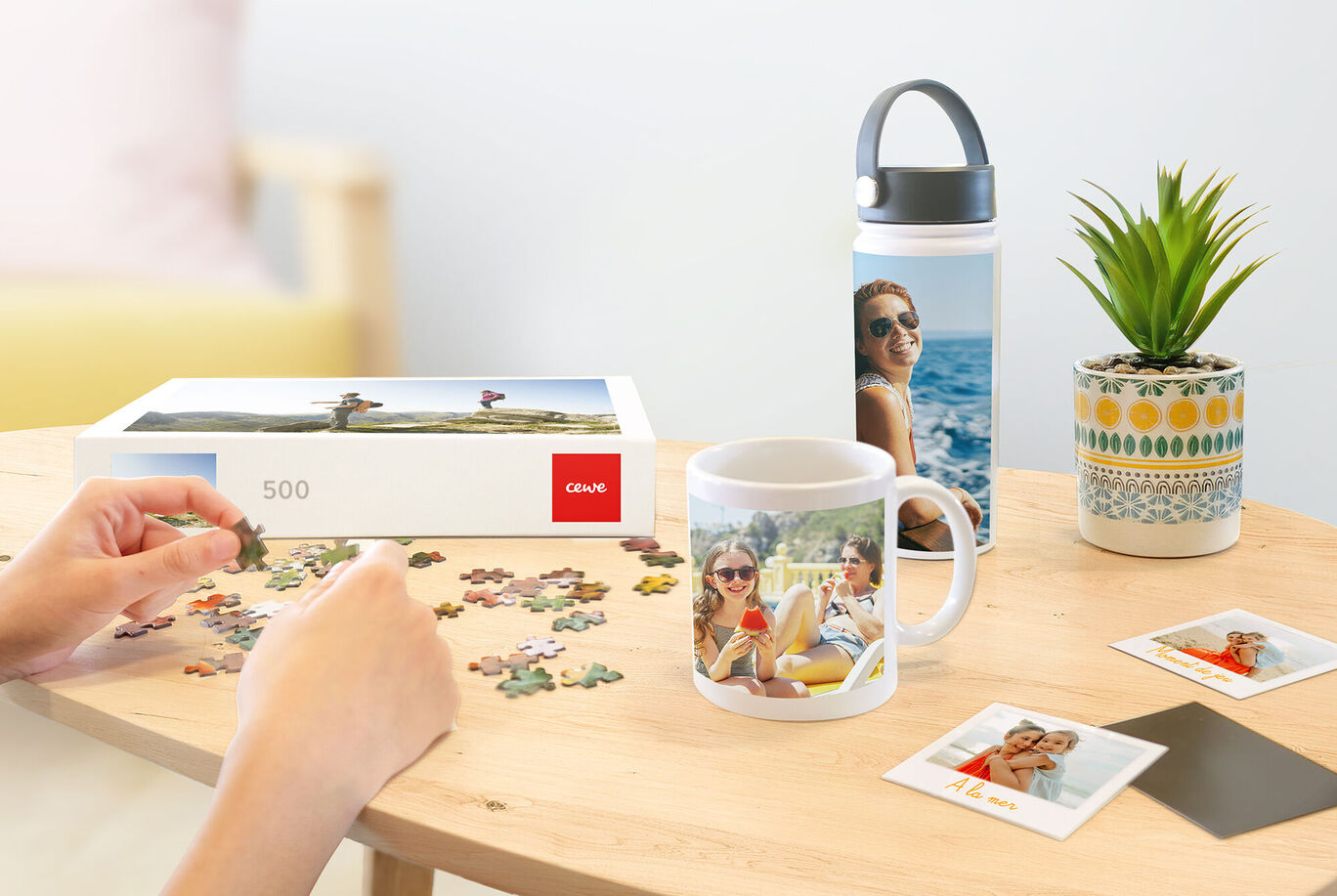 Una persona arma un rompecabezas CEWE. Al lado hay una taza y una botella de fotos. También hay una planta y fotos sobre la mesa.