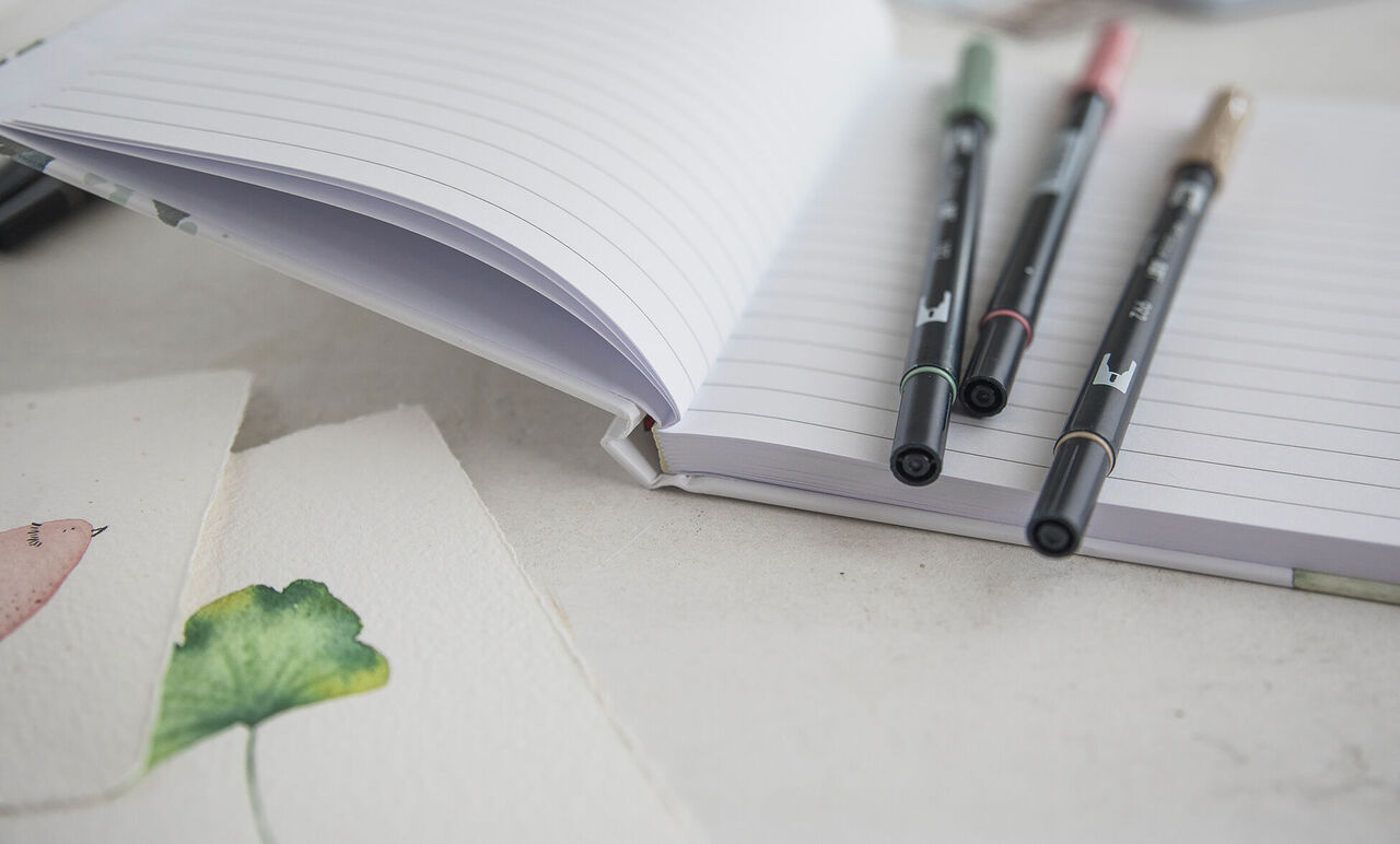 Cuaderno personalizado con líneas rectas en el interior y unos bolis para escribir.