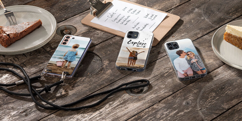 Tres fundas de móvil personalizadas sobre una mesa de madera. Una con una foto de un niño en la playa, otra con dos amigos celebrando y una con una pareja usando gafas de sol. Hay un bloc de notas y un pedazo de pastel en la mesa.