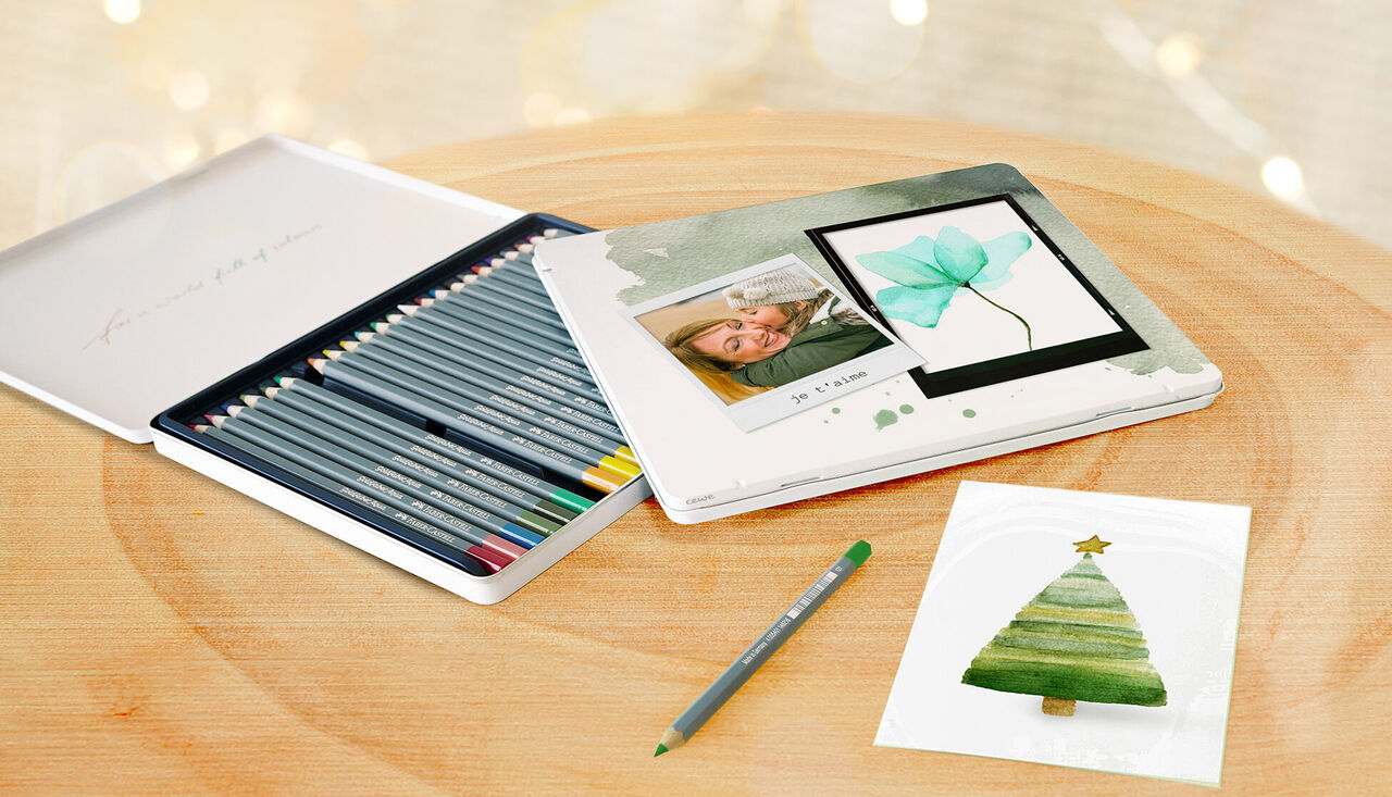 Una caja de regalo creativa con lápices de colores y una foto personal. Junto a ella, una tarjeta navideña hecha con acuarelas. Ideas de manualidades inspiradoras para momentos especiales.