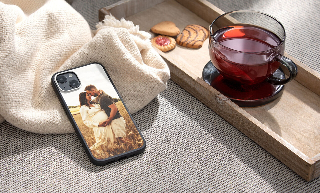 Un teléfono con una funda de fotografía que muestra a una pareja en un paisaje de pradera. Al lado, galletas y una taza de té.