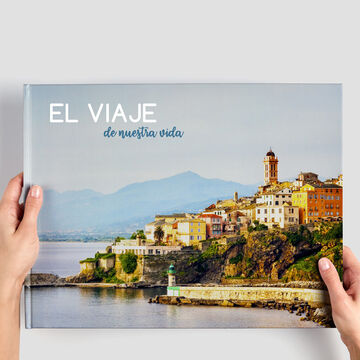 Una persona sostiene un fotolibro CEWE titulado 'El Viaje de nuestra vida' frente a un paisaje costero.