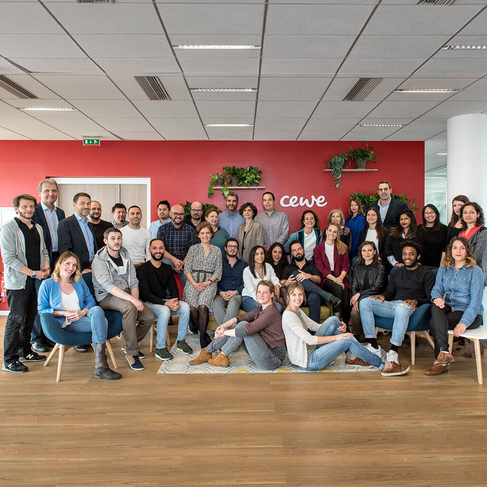 Un grupo de empleados se reunió feliz para una foto. Están de pie y sentados en una sala, rodeados de plantas y una pared roja con el logo de CEWE.