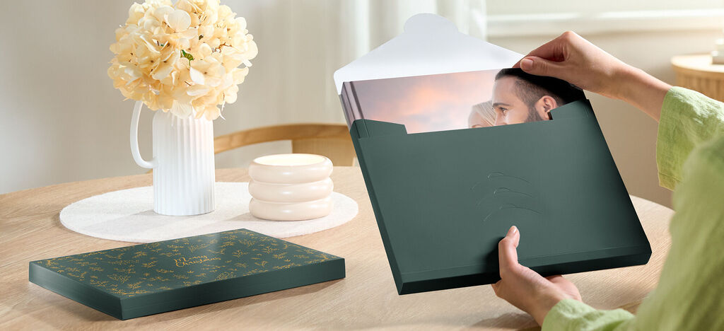 Una persona abre una bonita caja de regalo con un fotolibro. Al fondo hay un jarrón con flores y elementos decorativos.