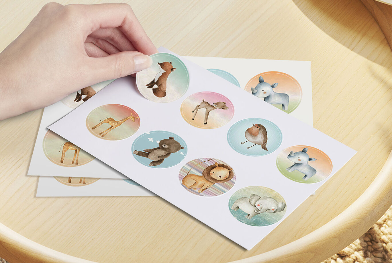 Una mano sostiene una pegatina de un animal adorable sobre una hoja con animales coloridos.