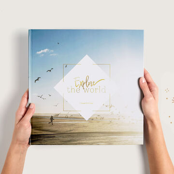 Una mano sostiene un álbum de fotos CEWE titulado 'Explora el Mundo', de fondo una playa.
