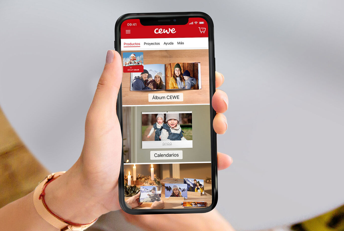 Una mano sostiene un smartphone con la app de CEWE Fotobuch. La app muestra un álbum y dos calendarios.
