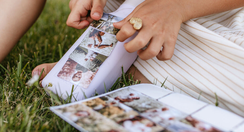 Una persona ve fotos en un libro fotográfico CEWE y sostiene otra imagen, rodeada de hierba verde.
