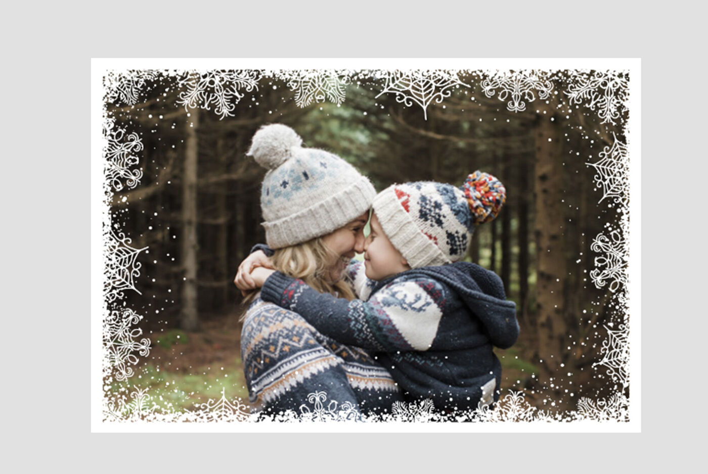 Madre lleva a su hijo en brazos, ambos con gorros de lana en un bosque invernal.