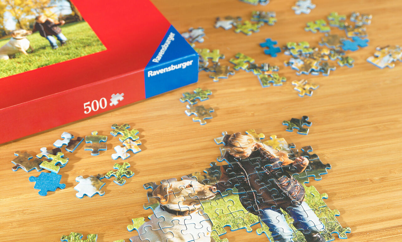 Un rompecabezas con imagen de dos niños y un perro. Piezas esparcidas en una mesa. La caja es roja y muestra la imagen a gran escala.